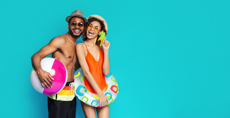 A happy black couple posing against a teal backdrop. The man is holding a pink and white beach ball while the woman is holding a colorful inflatable ring, copy space