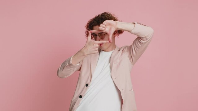 Caucasian man photographer gesturing picture frame with hands, looks through fingers and focusing on interesting moment, imitating zoom and cropping nice image. Young guy isolated on pink background