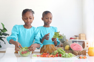Twin sister kid girl with curly hair braid African putting fresh vegetables and fruits after washing and cleaning in glass bowl for making salad. Cute children preparing salad healthy food at kitchen
