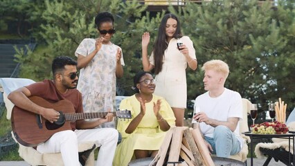 A group of diverse friends enjoys a summer picnic, with one of them playing a guitar while others sing along, drink wine