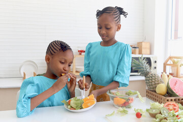 Twin sister kid girl with curly hair braid African eating fresh vegetables and fruits after washing and cleaning in glass bowl for making salad. Cute children preparing salad healthy food at kitchen.