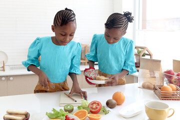 Twin sister kid girl with curly hair braid African taking down on recipes notebook during cooking at kitchen. Cute children with fresh ingredient, vegetable and fruits, to prepare healthy food at home