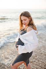 Pregnant woman 24 - 25 year old holding prenatal belly wearing stylish casual clothes over sea at beach outdoor. Maternity lifestyle.