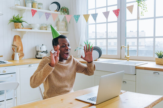 Excited African American man wearing a party hat celebrating his birthday online with laptop in festive kitchen setting. Black guy in festive hat congratulates friend on online video call on computer. - Powered by Adobe