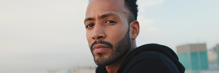Close-up, young bearded male athlete standing on the embankment, looking at the sea. Guy turns his...
