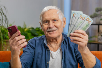 Planning family budget. Smiling senior man counting money cash use smartphone calculate domestic bills at home. Mature elderly grandfather satisfied of income, saves money for planned vacation gifts