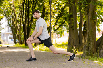 Fitness young man in sportswear doing lunges, and stretching legs on summer park. Warm up before jogging workout. Athletic male jogger training legs on footpath amidst trees. Squat lunges training.