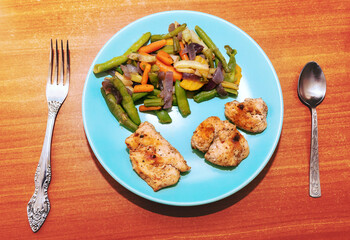 Blue plate with food including vegetables and meat. The plate has chicken, carrots, and green beans. Plate of food with a fork and spoon on a wooden table