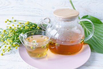 Herbal medicinal tea with chamomile flowers in a glass teapot, a bouquet of fresh flowers, on a wooden light background.