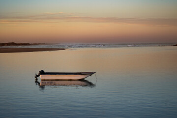 A small boat floats peacefully on calm water at sunset, with a vibrant orange sky reflecting off the water's surface and a distant shoreline silhouetted against the horizon.