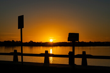 A stunning sunset over a calm lake with silhouettes of signs and a railing in the foreground. The sun's golden reflection shimmers on the water, creating a serene and picturesque scene.
