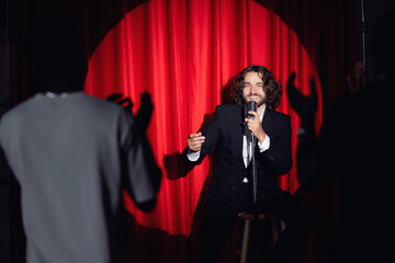 Smiling man speaking to microphone performing in comedy show open mike stand up on stage with red...