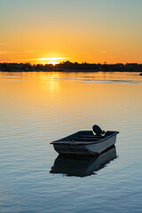 A small boat floats peacefully on calm water at sunset, with a vibrant orange sky reflecting off the water's surface and a distant shoreline silhouetted against the horizon.