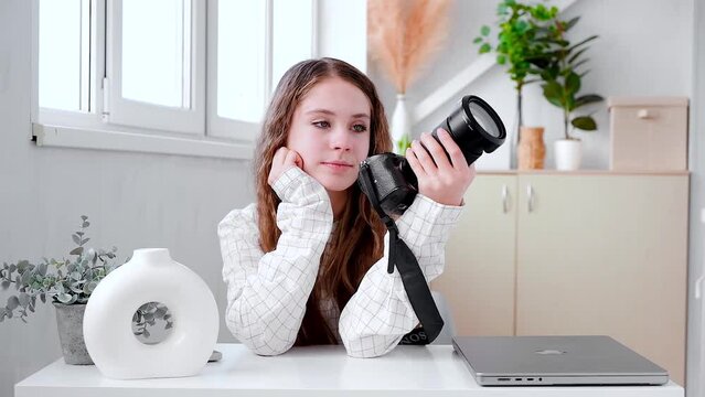 A beautiful young photographer girl holds a camera in her hands while sitting at a table in a modern white interior in a bright room. Samara, Russia - 23 May 2024