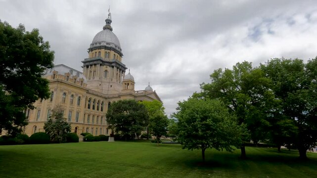 Long view of the Illinois State Capitol Building in Springfield, IL, USA. Cloudy overcast skies overhead. Springtime scene with a lush green lawn and full trees.