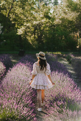 Beautiful young woman, happy girl in a straw hat, summer sundress dress walks in a lavender purple field at sunset outdoors. Photography, portrait, lifestyle.