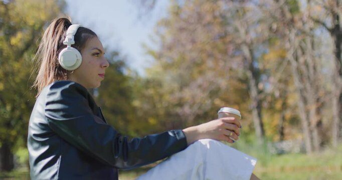 Beautiful girl with headphones holding coffee cup. Fashionable young lady sitting in the park.