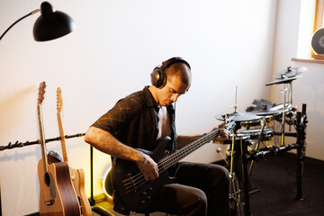 Fototapeta premium Young musician in headphones playing bass guitar near music instruments in studio