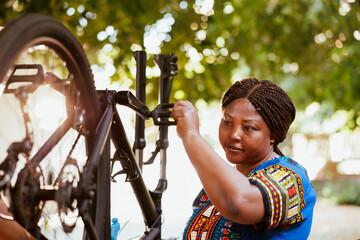 Naklejka premium Young woman outdoors repairing bicycle tire with professional equipment. African american woman holding and checking bike wheel in yard for maintenance.
