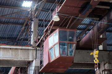 Overhead crane with a linear cross-beam and a yellow hook in the shop of the machine-building plant. Crane operator's cabin and trolley of the boom crane. Ladder to the roof through the hatch