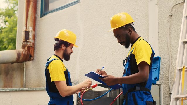 Seasoned mechanic doing condenser investigations, filter replacements and necessary fixes to prevent HVAC system failures while adept repairman coworker writes findings on clipboard
