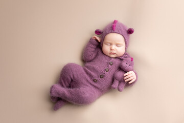 Little adorable newborn baby girl sleeping in a hat with dragon ears. Neutral background. Studio shot of a ten-day-old newborn baby. Beginning of life. Fairy tale. Dragon toy