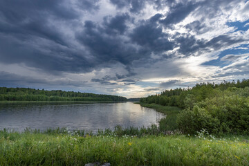 A picturesque view of a lake surrounded by lush greenery, with a dramatic sky featuring thick, grey clouds overhead. The sun peeks through the clouds, casting a warm glow on the water.