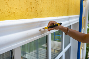 A hand is holding a utility knife and is removing excess caulk from a white trim above a window on...
