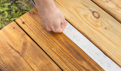 A hand carefully presses down white tape on a freshly stained wooden deck, ensuring a clean line for the next step in the project.
