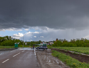 A dark, stormy sky looms over a rural road. A car has pulled over, waiting for the rain to pass. The road is wet and the trees lining the road are green and lush..