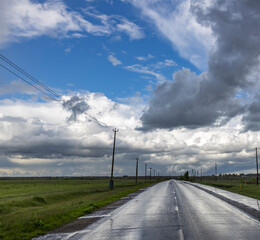 Fototapeta premium A paved road stretches into the distance, reflecting the sky, which is covered in large, dramatic clouds after a recent rain..