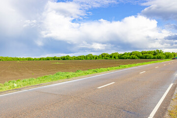 An empty asphalt road stretches into the distance, bordered by a field of brown earth and a line of verdant trees. Above, fluffy white clouds drift across a bright blue sky..