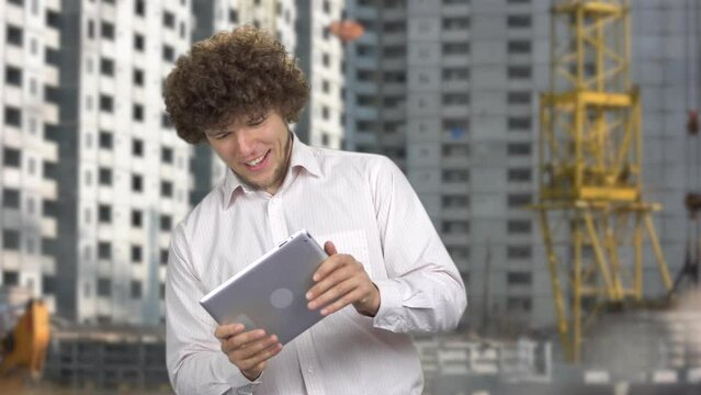 Happy excited man with curly hair playing a video game on the tablet pc device. Construction site in the background.