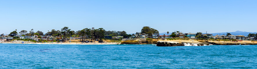 Fototapeta premium Scenic panoramic view of Santa Cruz shoreline as seen from the water, featuring sandy beaches, residential homes, and tall trees against a bright, clear sky
