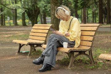 In Yoyogi Park, Shibuya, Tokyo, a Japanese man in his twenties wearing yellow clothes, sitting on a secluded bench, operating a smartphone along with headphones, music, and coffee.