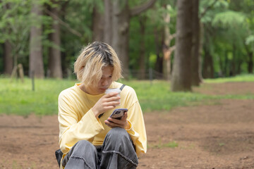 In early summer, in a quiet area of Yoyogi Park, Shibuya, Tokyo, a Japanese man in his twenties wearing yellow clothes, sitting on a tree stump, drinking coffee and operating a smartphone.