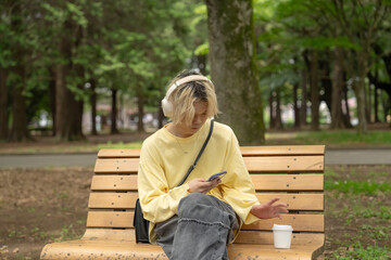 In Yoyogi Park, Shibuya, Tokyo, a Japanese man in his twenties wearing yellow clothes, sitting on a secluded bench, operating a smartphone along with headphones, music, and coffee.