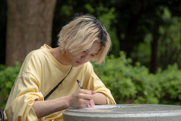 In early summer, a young Japanese man in his early twenties wearing yellow clothes, sitting at a table in Yoyogi Park, Shibuya, Tokyo, writing on paper during the day.