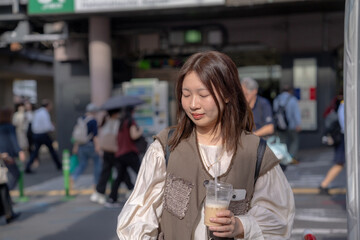 In early summer, a young Chinese exchange student in her twenties, standing in front of the busy Hamamatsucho Station in Minato, Tokyo, holding an iced café au lait in a plastic cup, 