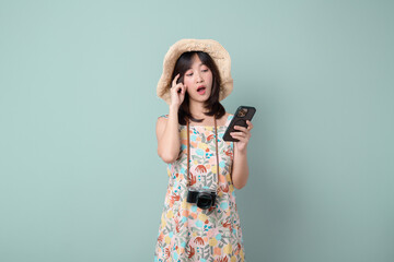 Thinking on vacation concept. Happy Asian woman wearing casual summer dress with hat and camera thinking gesture while using mobile phone isolated on pastel green background.