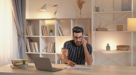 Phone consultation. Busy worker. Check report data. Business man talking on cellphone about information in documents sitting at desk with laptop in home office interior.