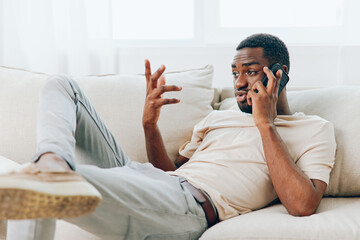 Happy African American man sitting on a black sofa, holding a smartphone and smiling while making a video call The modern apartment provides a comfortable background for the relaxed chat, showcasing