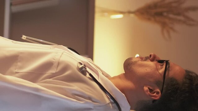 Vertical video. Doctor relaxing. Coffee break. Enjoying moment. Dreamy calm smiling medical worker man drinking hot beverage from cup resting in clinic interior.