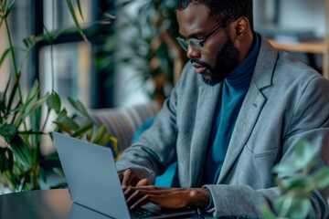 man wearing glasses is sitting at a table with a laptop in front of him. He is focused on his work, possibly typing or browsing the internet. Concept of productivity and concentration