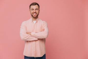 Cute unshaven man smiling with healthy teeth and crossed arms in pink shirt posing isolated on pink...