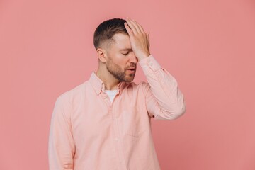 Cute unshaven man in pink shirt suffering from headache and holding hand to head isolated on pink...