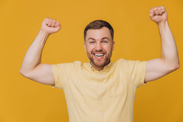 Winner! Cute unshaven man in yellow shirt waving fists in joy smiling and posing isolated on yellow background with copy space