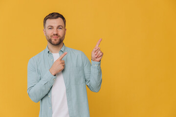 Cute happy unshaven man in mint shirt showing fingers up at copy space isolated on yellow background