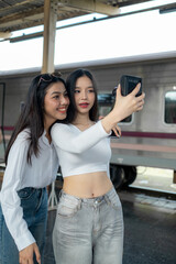 Smiling women on the station platform with happily looking into a smartphone, girls standing at train station take selfie photo on smartphone