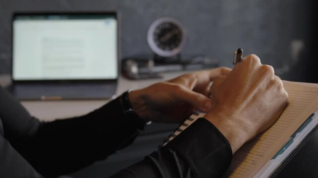 Close-up view of a man writing lyrics in a notebook with a laptop in the background.